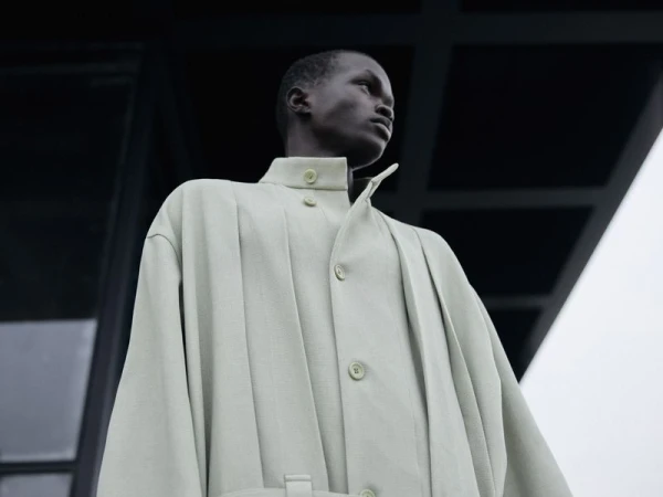 Low-angle shot of a black male model in a light green structured coat against a dark architectural exterior.