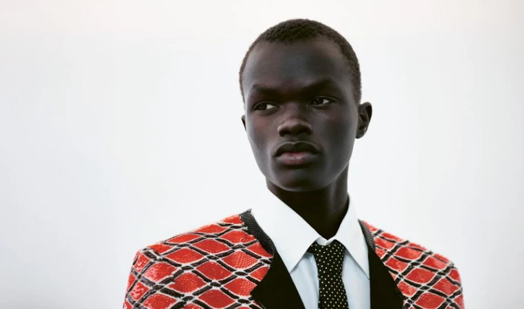 Portrait of a black male model wearing a patterned red jacket, white collared shirt, and black polka dot tie against a white background.
