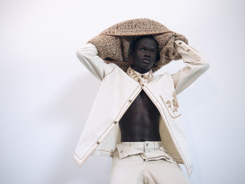 Black male model holding a textured brown woven garment above his head while wearing an off-white jacket and trousers.