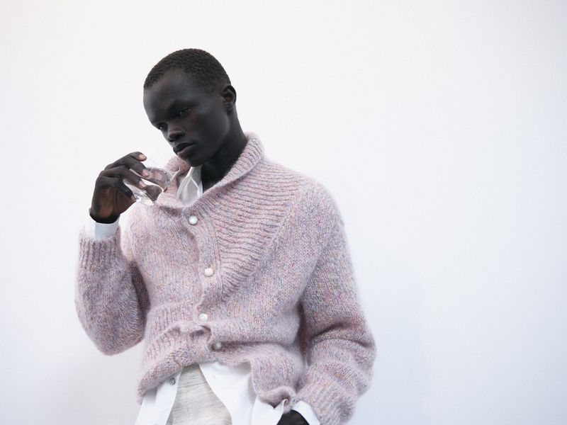 Black man holding a glass of water wearing a pink knit cardigan against a white background