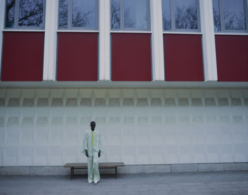 Model standing in front of a textured light grey building wall and red panels wearing a light patterned suit