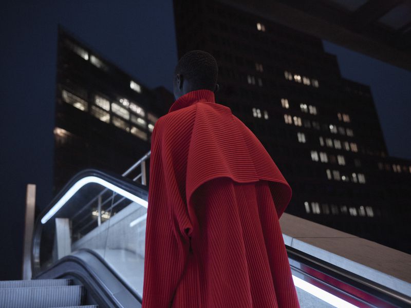 Model seen from behind wearing a red ribbed cape coat on an escalator at night with dark buildings in the background.