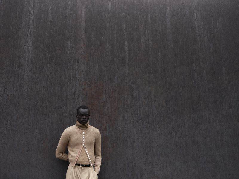 Black male model wearing a beige button-up sweater standing against a dark textured metal wall.