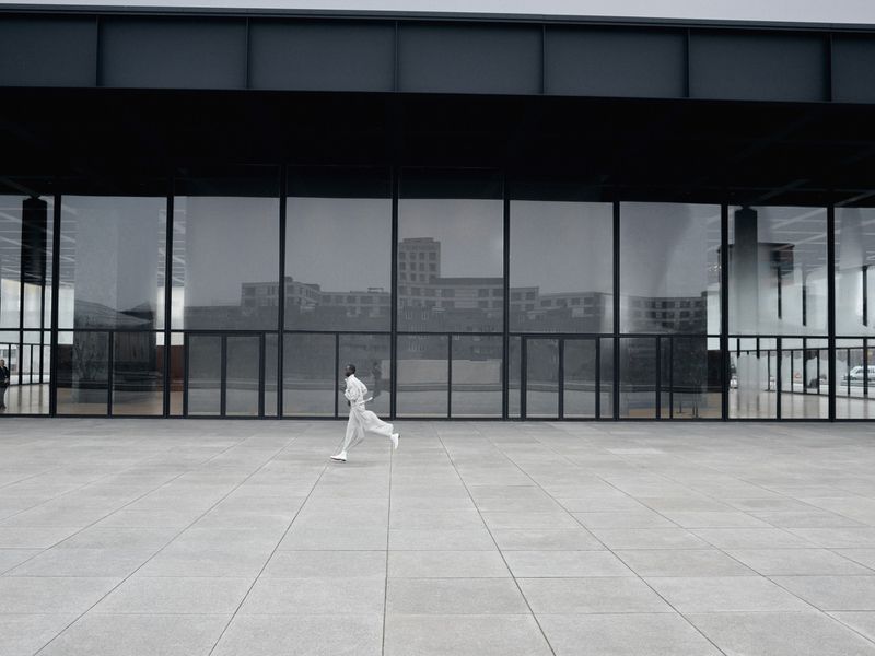 Model in white clothing running across a large grey concrete plaza in front of a modern glass and dark metal building