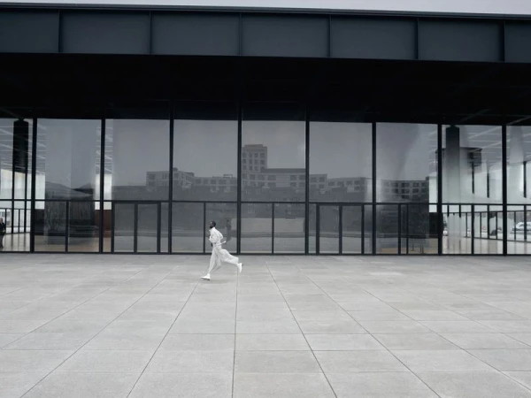Model in white clothing running across a large grey concrete plaza in front of a modern glass and dark metal building