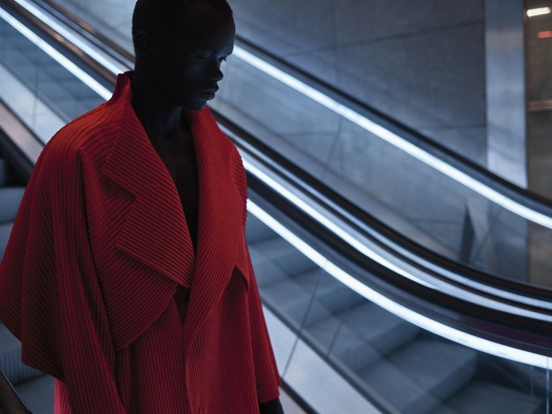 Model wearing a pleated red coat standing on an escalator with glowing illuminated handrails.