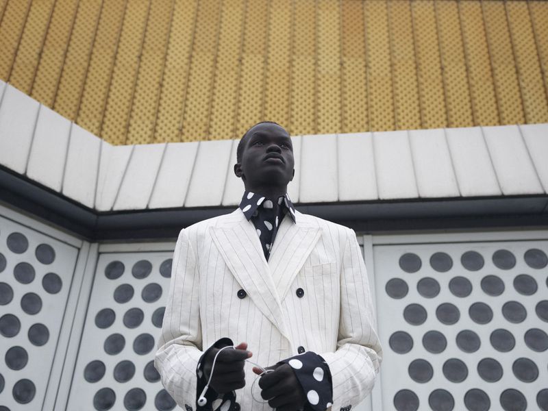 Model wearing a white pinstripe double breasted jacket and polka dot shirt standing against an exterior wall.