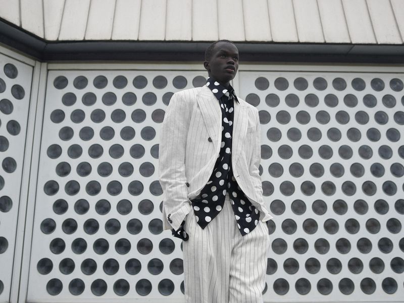 Black man wearing a white pinstripe suit and black polka dot shirt standing outdoors against a patterned architectural wall.