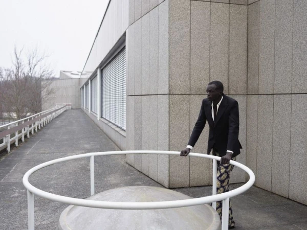 Model Wal Ruop in a dark blazer and patterned trousers leaning on a white railing outdoors