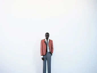 Model standing against a plain white wall wearing a patterned orange blazer and grey trousers.