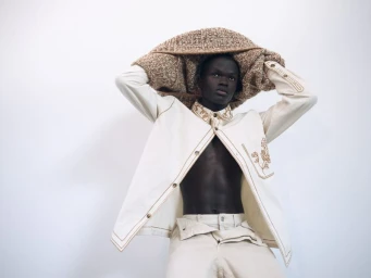 Black male model holding a textured brown woven garment above his head while wearing an off-white jacket and trousers.
