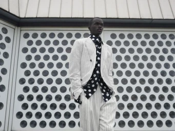 Black man wearing a white pinstripe suit and black polka dot shirt standing outdoors against a patterned architectural wall.