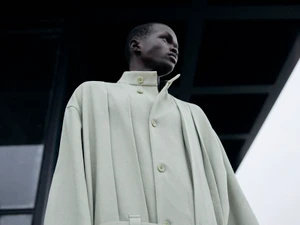 Low-angle shot of a black male model in a light green structured coat against a dark architectural exterior.