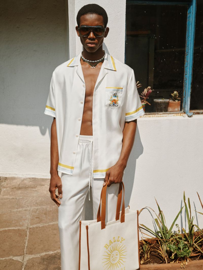 Model wearing a white shirt and trousers holding a branded tote bag outside a white building