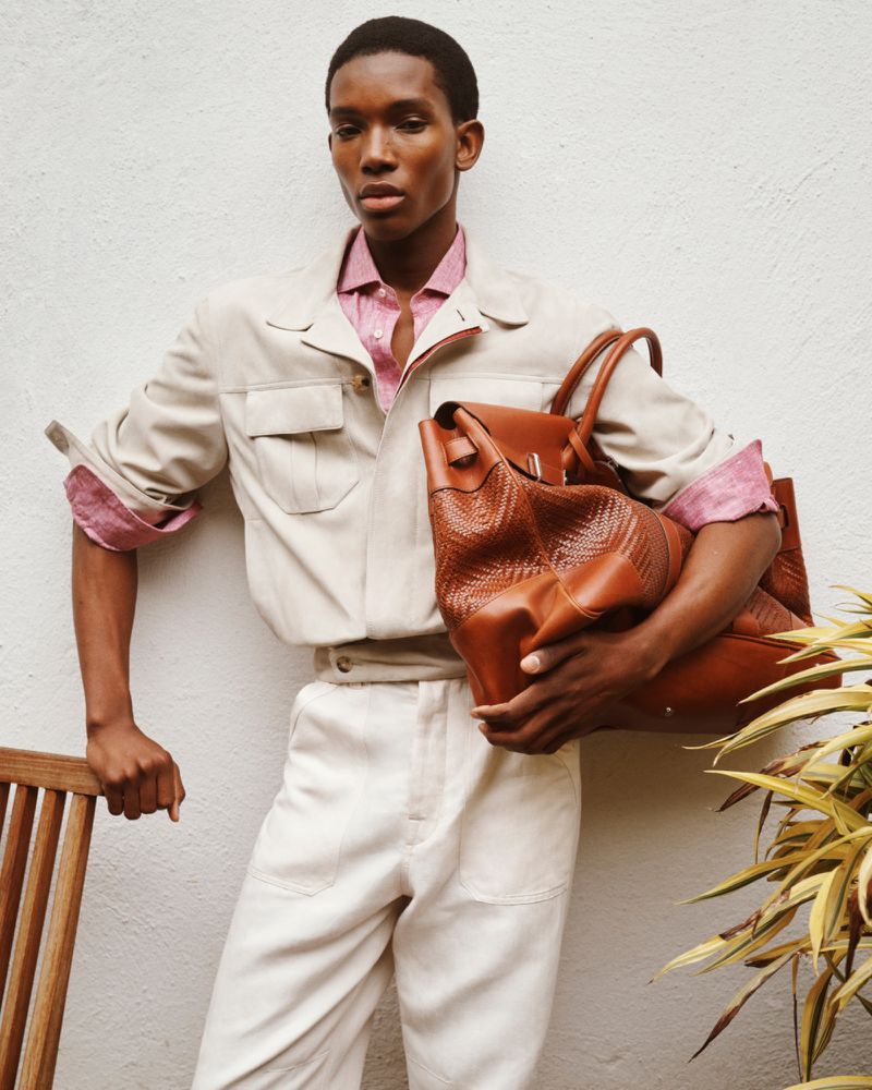 Model holding a large leather bag wearing an off-white jacket and pink shirt against a textured white wall.