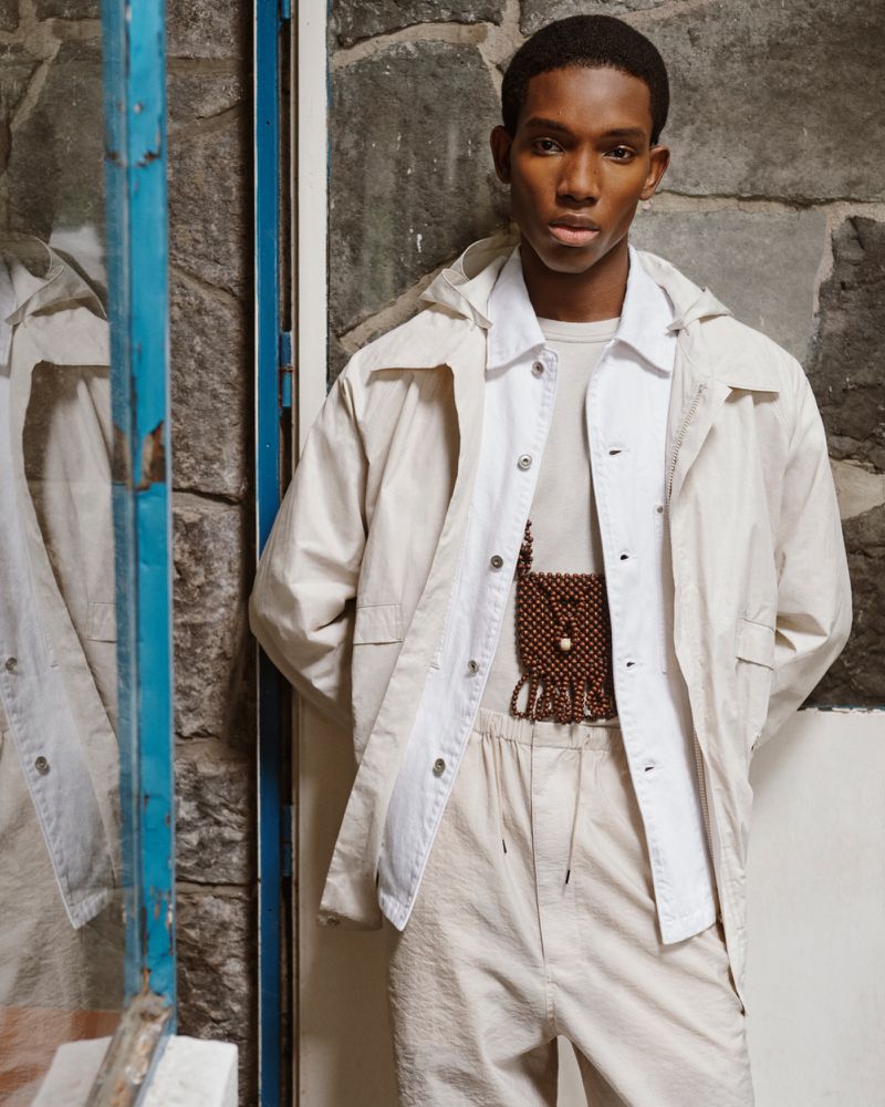 An African American man poses in a casual beige outfit near a stone wall and blue-framed door, showcasing fashion and style.