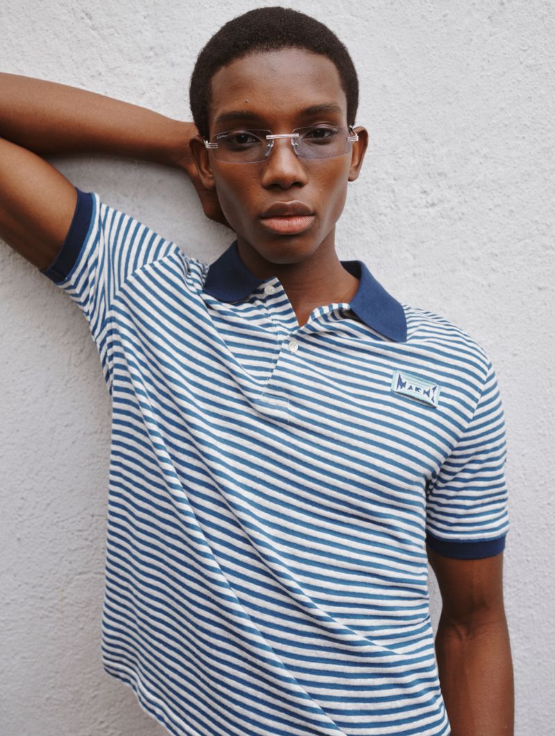 Model Ibra Niakate wearing a blue and white striped polo shirt against a textured white wall for Farfetch