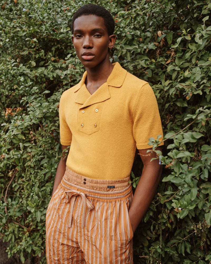 Model wearing a yellow short-sleeve polo shirt and striped trousers standing outdoors in front of leafy foliage.