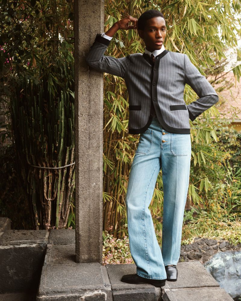 Model leaning against a concrete pillar in a pinstripe jacket and light denim jeans amidst lush bamboo foliage.