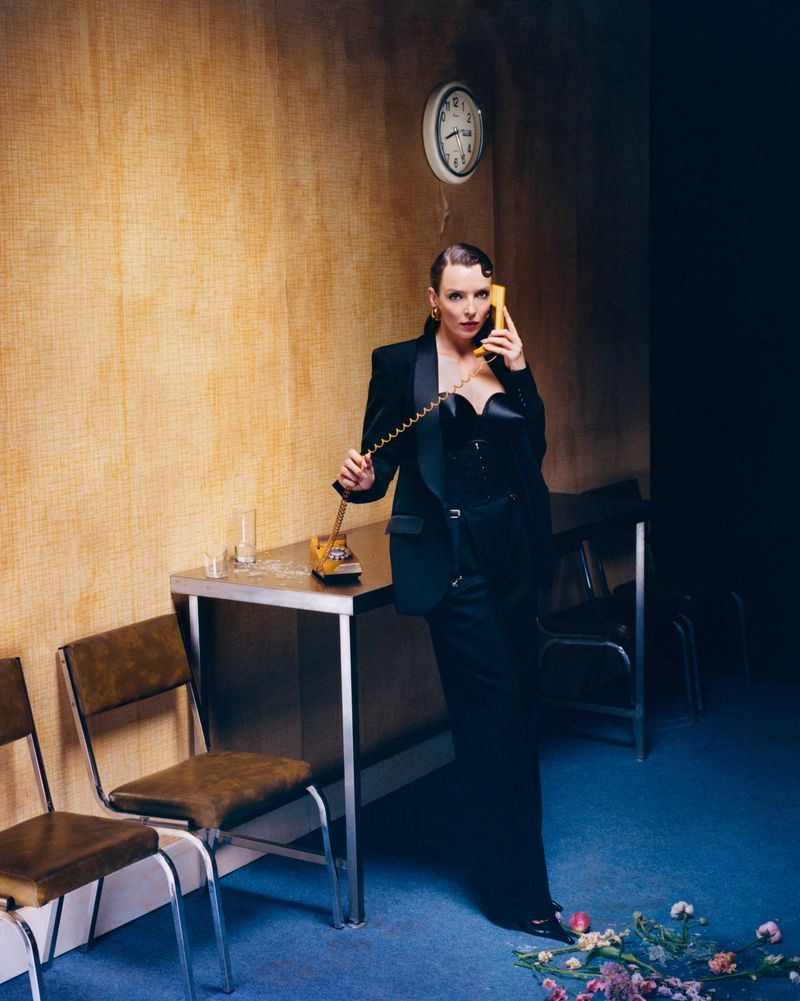 Woman in a black suit answers a yellow phone in an odd room with a clock, table, and flowers on the blue carpet.