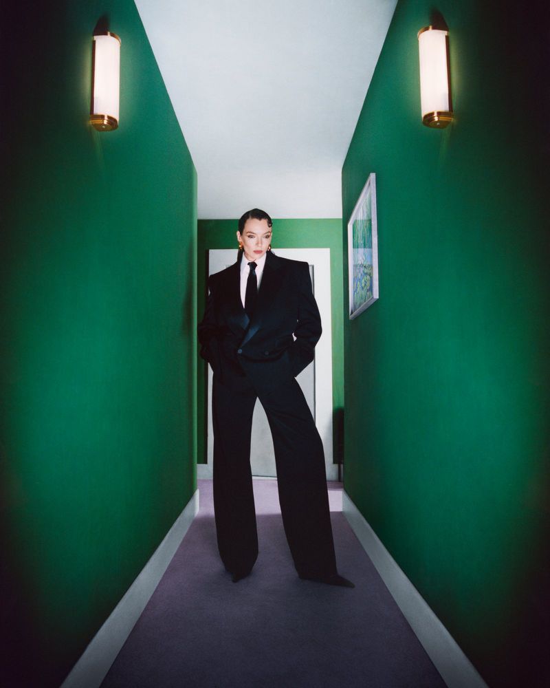 A person in a black suit stands in a green hallway with wall lamps. The photo features a corridor with a painting on the wall.