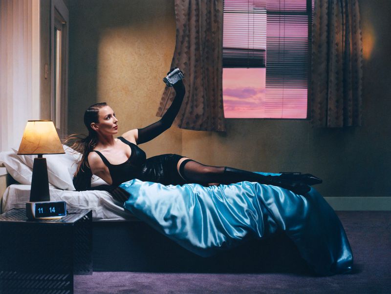 Adult woman in black lingerie and gloves lying on satin bed with a vintage camera, bedroom window, and digital clock in soft light.