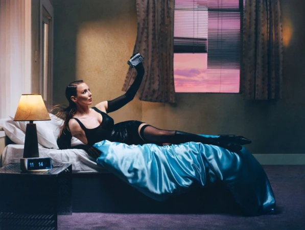 Adult woman in black lingerie and gloves lying on satin bed with a vintage camera, bedroom window, and digital clock in soft light.