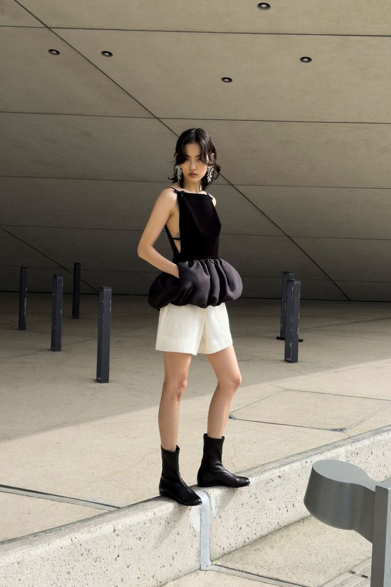 Model in a black sleeveless peplum top and white shorts standing outdoors against a concrete architectural backdrop