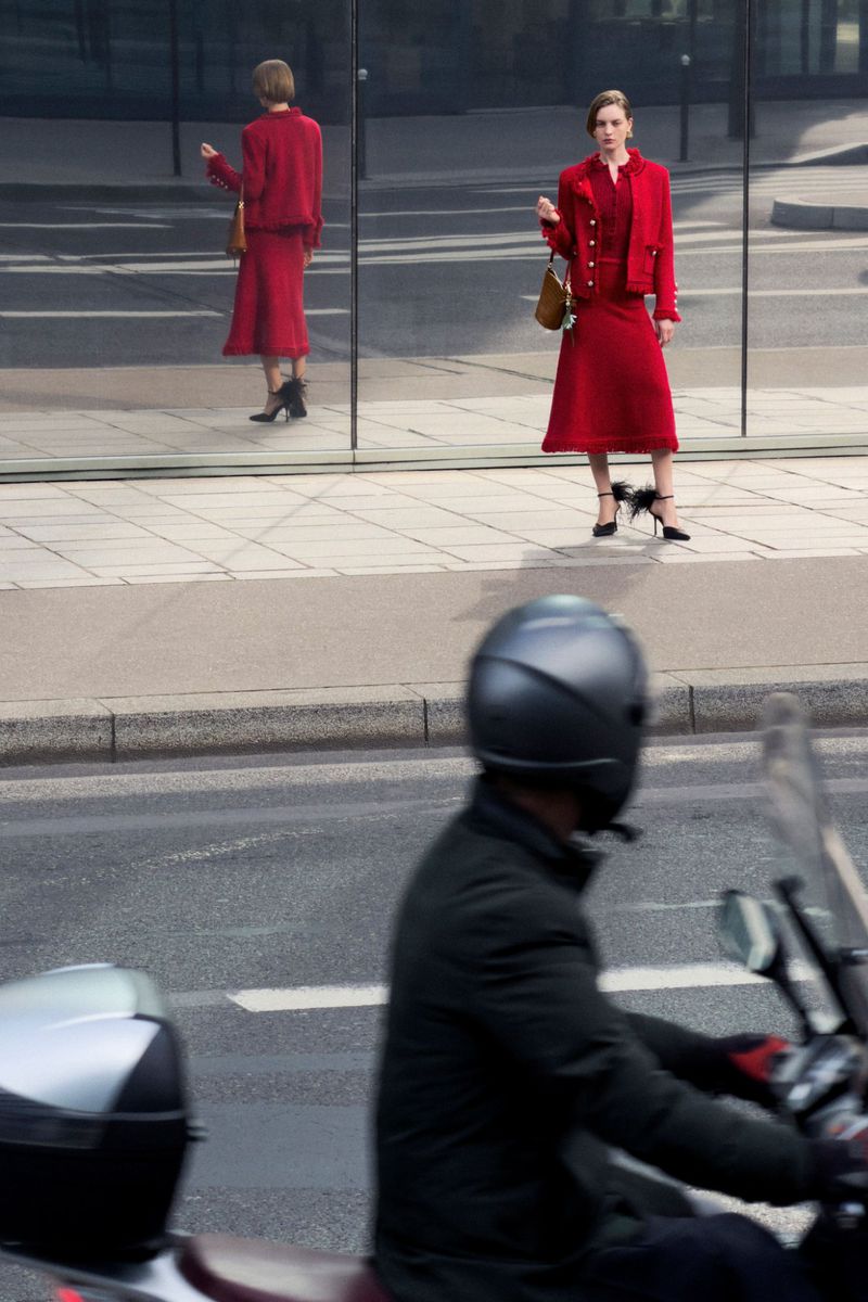 Model in a red skirt suit holding a brown handbag standing on a paved sidewalk next to a large glass storefront reflection.