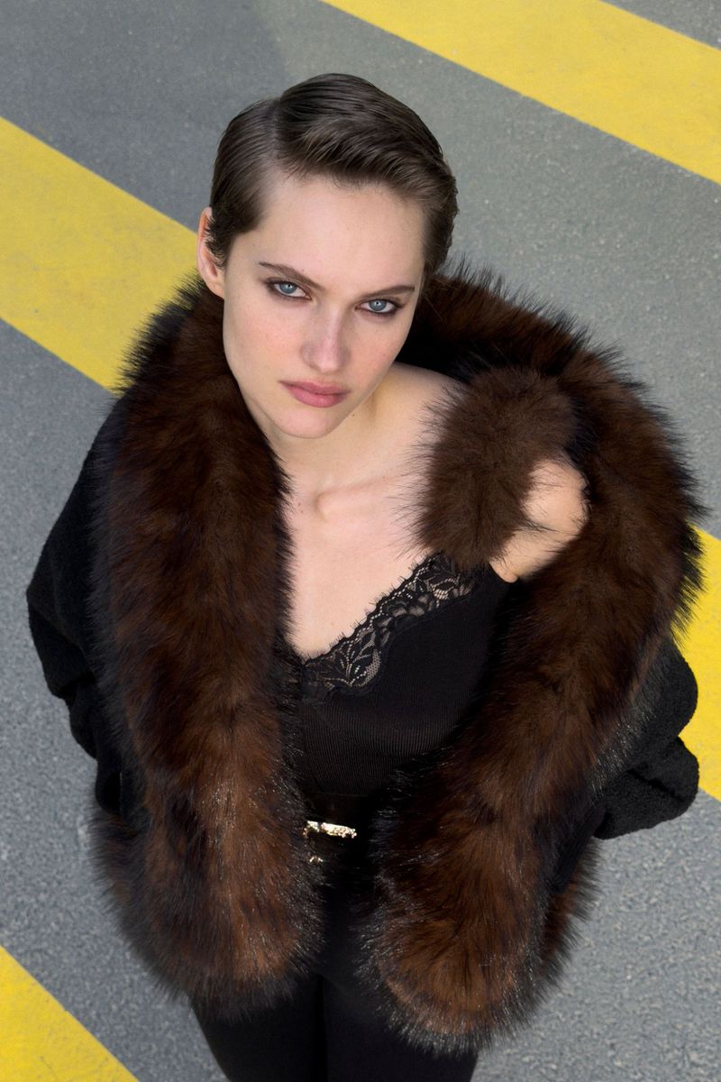 Model in a black top with a brown furry collar looking up against a grey asphalt background with yellow lines.
