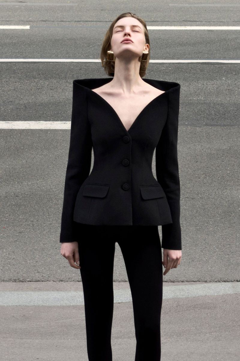 Model wearing a structured black blazer with prominent square shoulders standing outdoors on asphalt pavement.