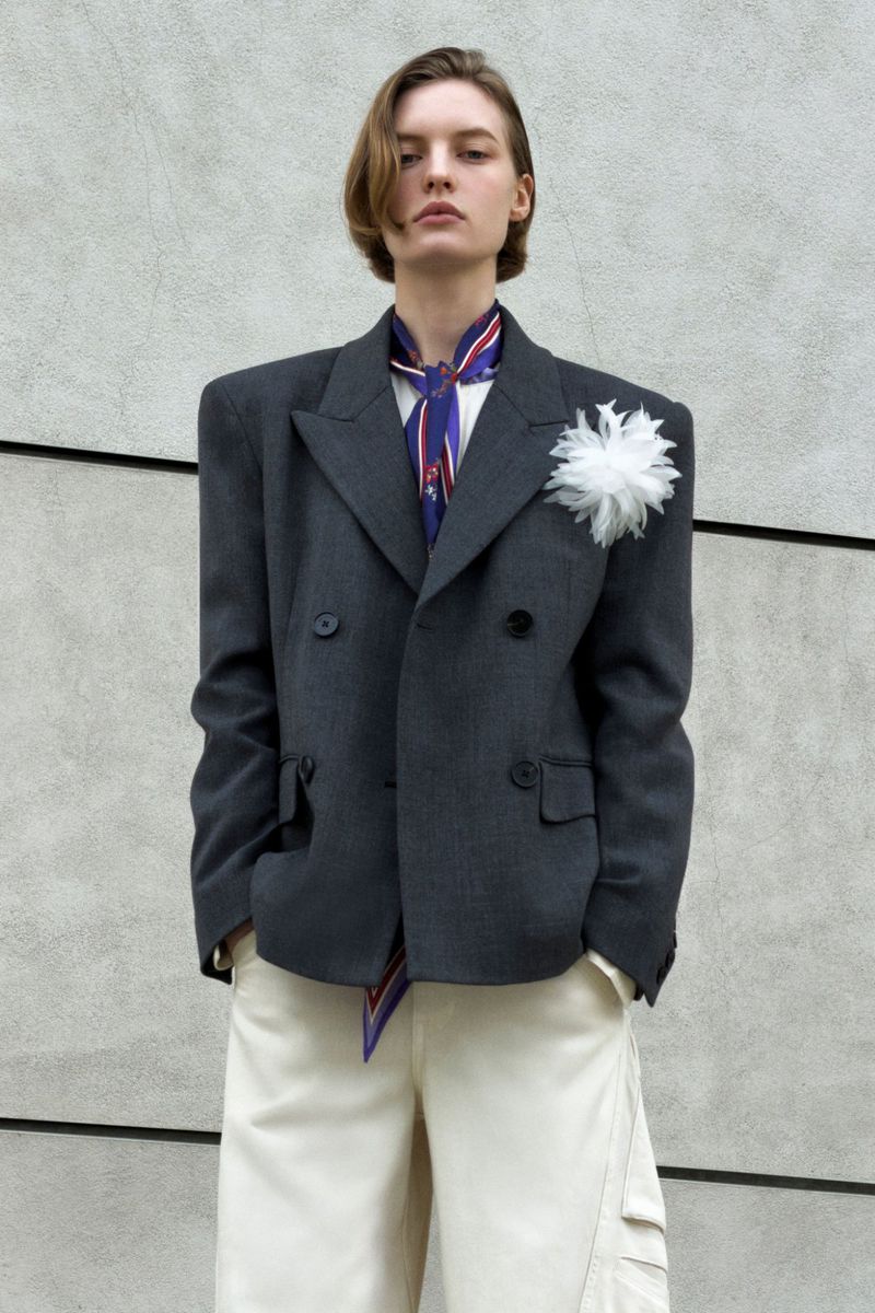 Model wearing a structured grey double-breasted blazer and white trousers against a concrete wall