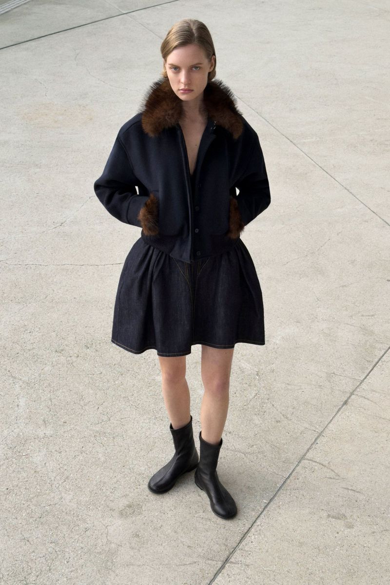 Full-length shot of a young woman wearing a dark coat with fur trim and a dark skirt with black boots, standing on a concrete surface.