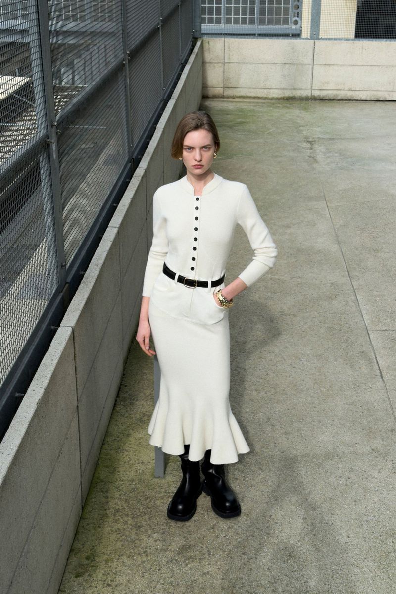 Model wearing a cream knit cardigan and matching skirt with a black belt on a concrete rooftop