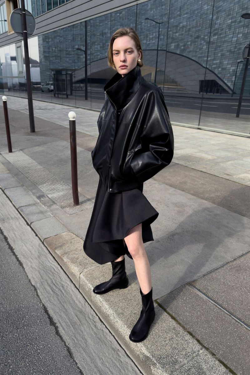 Model in a black leather jacket and black dress standing on a concrete street outdoors.