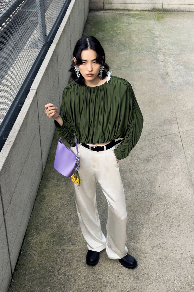 Model in olive green pleated top and white trousers holding a light purple handbag on concrete stairs