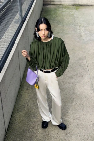 Model in olive green pleated top and white trousers holding a light purple handbag on concrete stairs