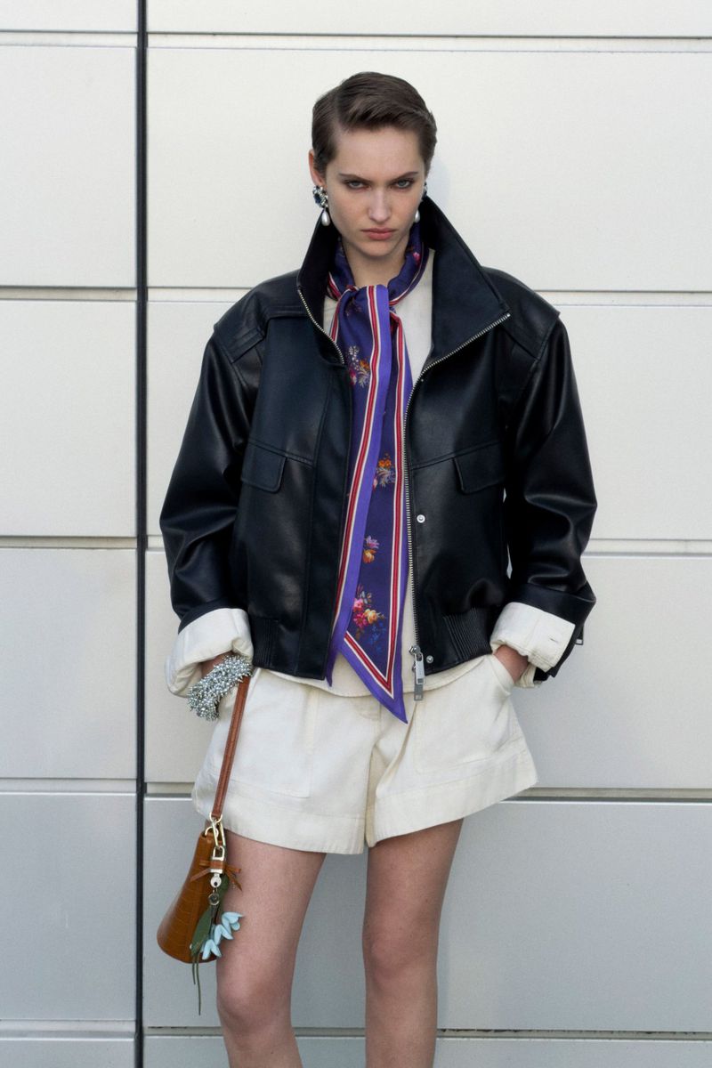 Model wearing a black leather jacket and white shorts with a purple patterned neck scarf standing against a tiled exterior wall