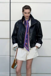 Model wearing a black leather jacket and white shorts with a purple patterned neck scarf standing against a tiled exterior wall