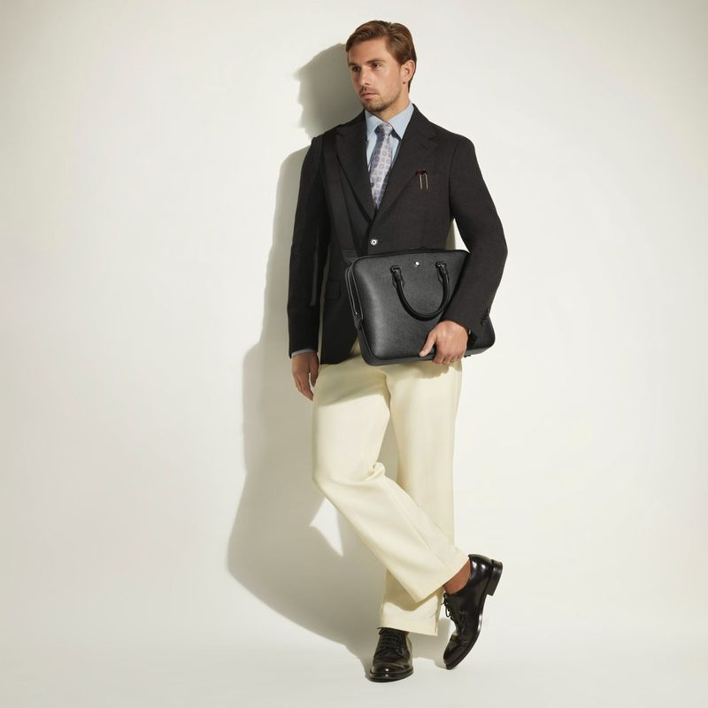 Model wearing a dark blazer and cream trousers holding a black leather briefcase against a white seamless background