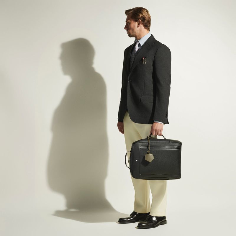 Adult man in a dark blazer stands against a white background holding a black briefcase, casting a soft shadow to the left.