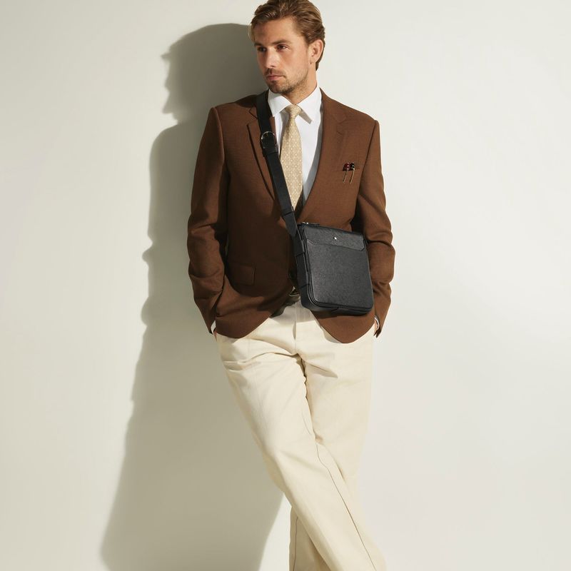 Model wearing a brown blazer, white shirt, and light trousers with a black leather crossbody bag against a white wall.