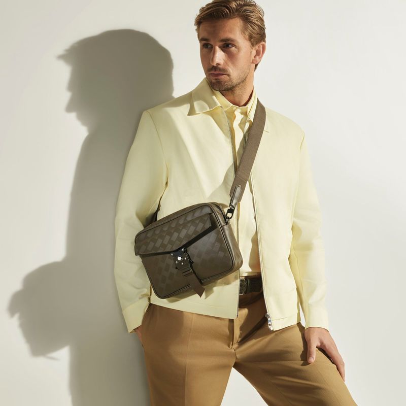 Man wearing a light yellow jacket and tan trousers with a dark crossbody bag against a white wall.