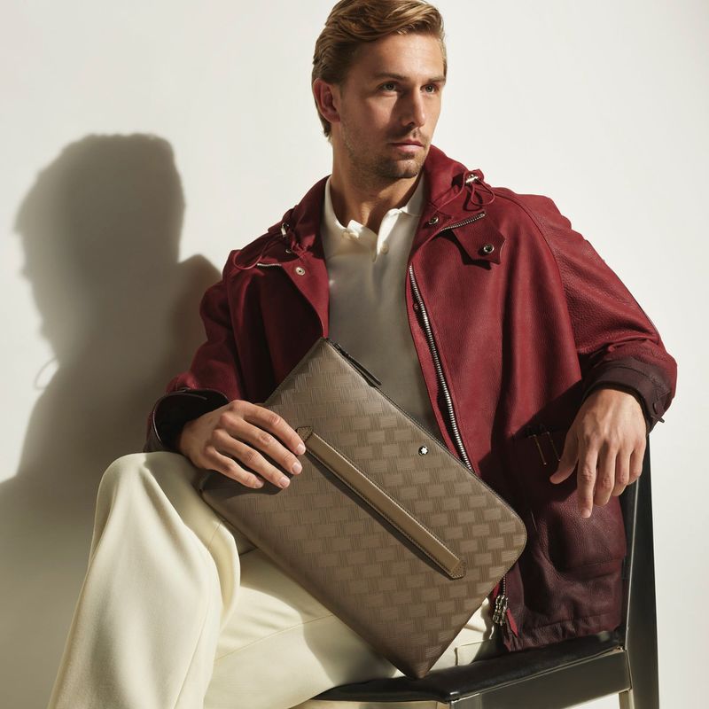Seated male model in burgundy jacket holding patterned leather folio bag against plain white background