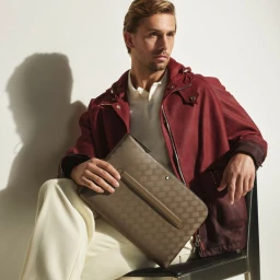 Seated male model in burgundy jacket holding patterned leather folio bag against plain white background