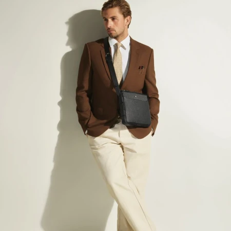 Model wearing a brown blazer, white shirt, and light trousers with a black leather crossbody bag against a white wall.