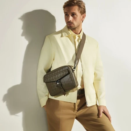Man wearing a light yellow jacket and tan trousers with a dark crossbody bag against a white wall.