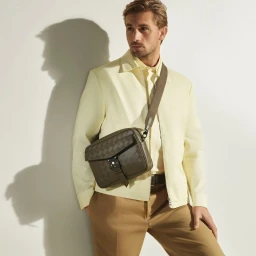 Man wearing a light yellow jacket and tan trousers with a dark crossbody bag against a white wall.