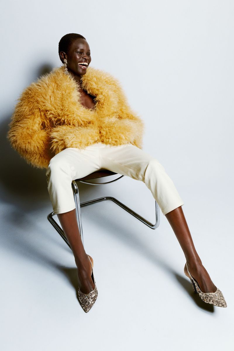 Black model laughing while seated in a metal chair wearing a fluffy yellow jacket, white pants, and heels.