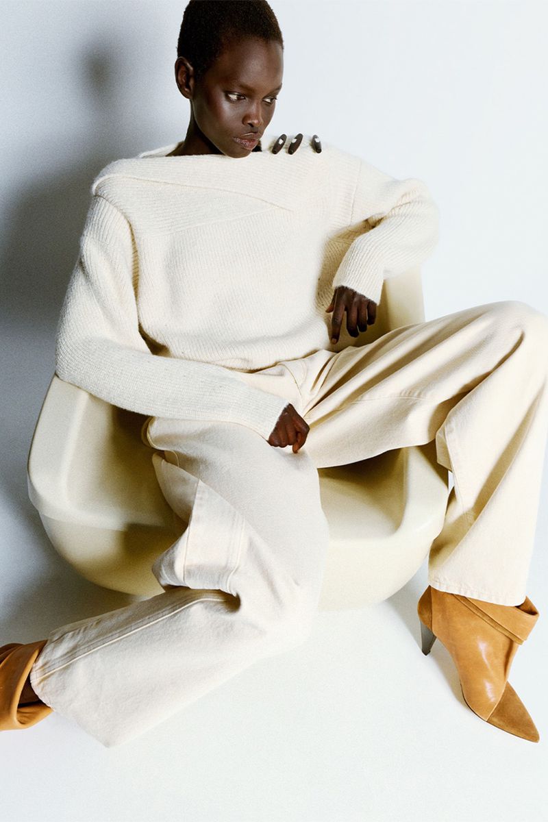 Model with short hair wearing a cream knit sweater and tan boots seated in a chair against a white background